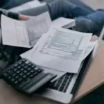 Person prüft Abrechnungsunterlagen mit Taschenrechner auf einem Laptop