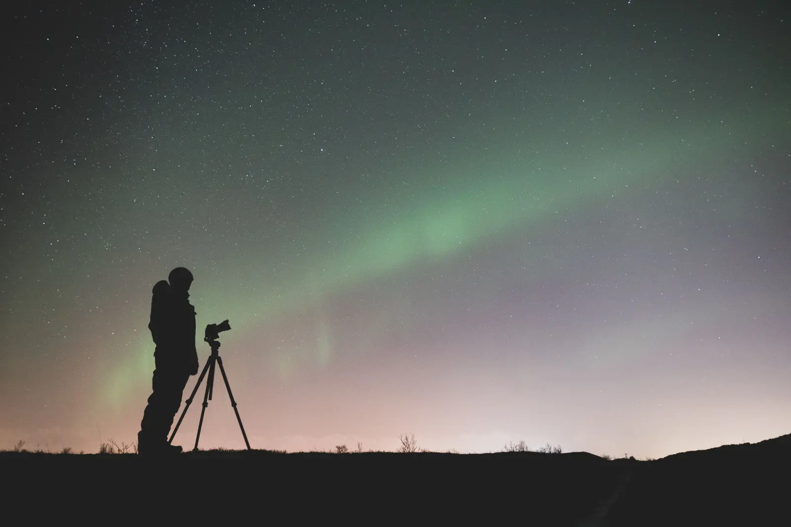 Silhouette einer Person mit Kamera auf einem Stativ unter einem schwachen grünen Polarlicht