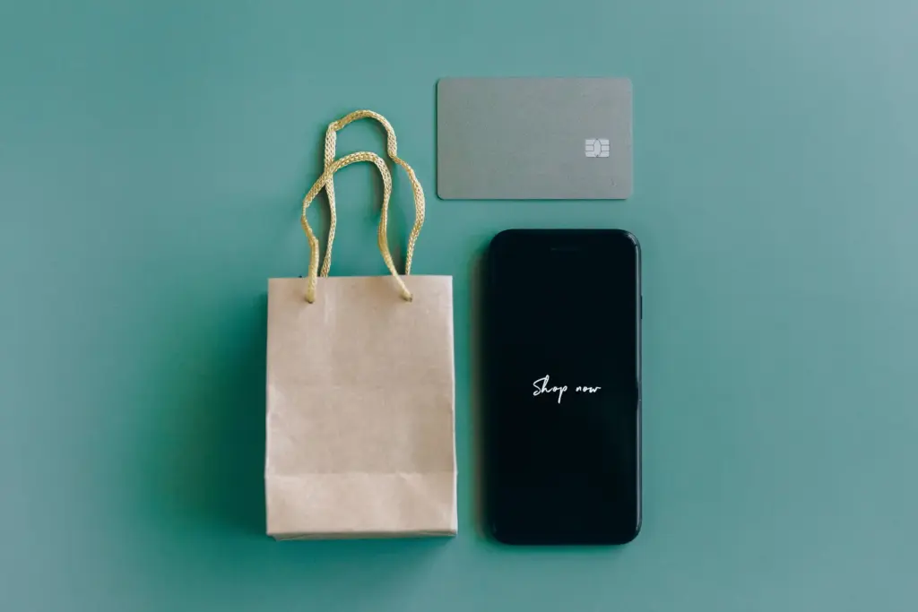 Smartphone mit der Aufschrift Shop now neben Einkaufstasche und Bankkarte