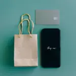 Smartphone mit der Aufschrift Shop now neben Einkaufstasche und Bankkarte