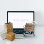Laptop mit Online-Shop sowie Pakete und kleiner Einkaufswagen auf einem Tisch