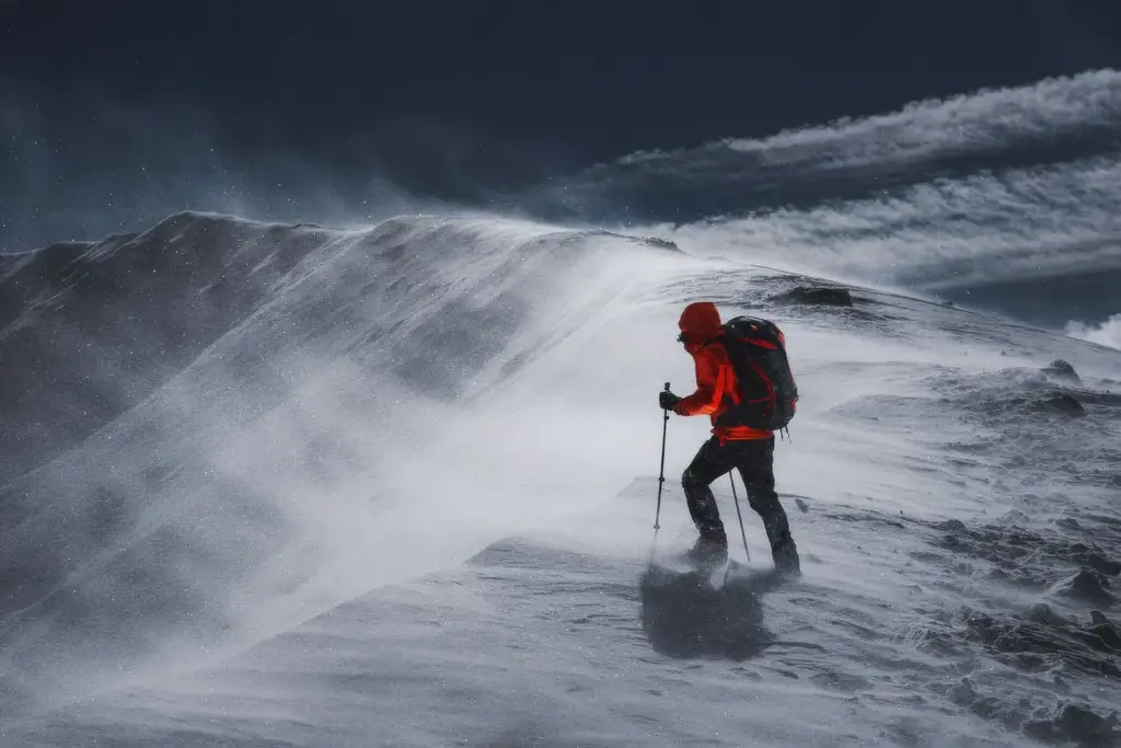 Wanderer in roter Jacke bei starkem Wind und Schneetreiben auf einem winterlichen Bergrücken