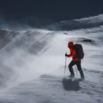 Wanderer in roter Jacke bei starkem Wind und Schneetreiben auf einem winterlichen Bergrücken